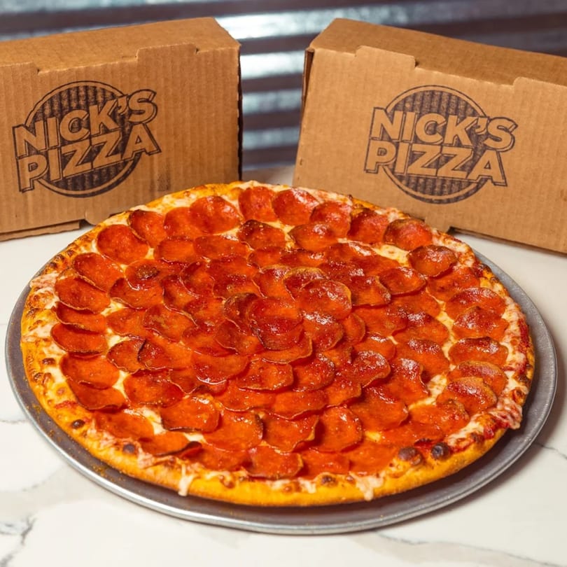 A large pepperoni pizza on a metal pan with two Nick's Pizza boxes in the background.